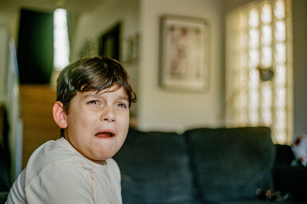 A child sitting on a dark couch in a living room with natural lighting.