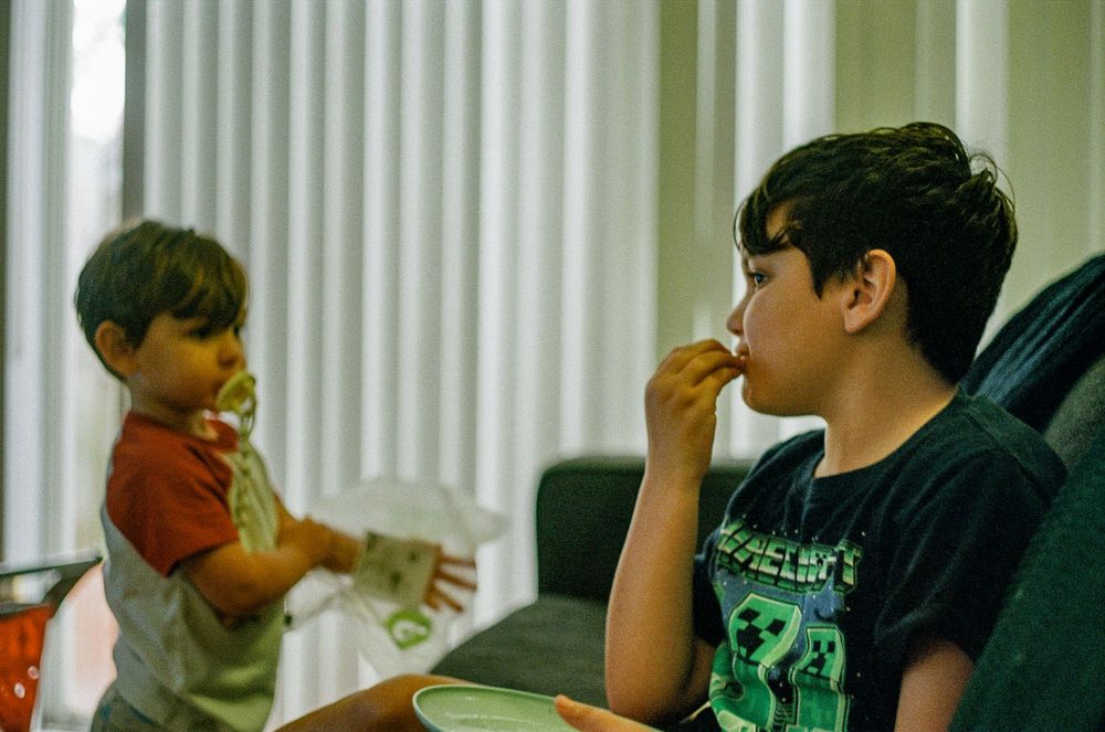 Two children interacting while having snacks in a room with vertical blinds.