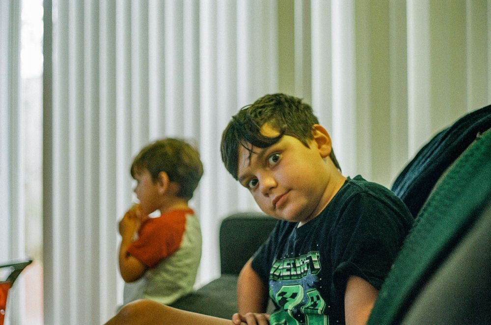 A child in a Minecraft t-shirt sitting on a chair near vertical blinds.