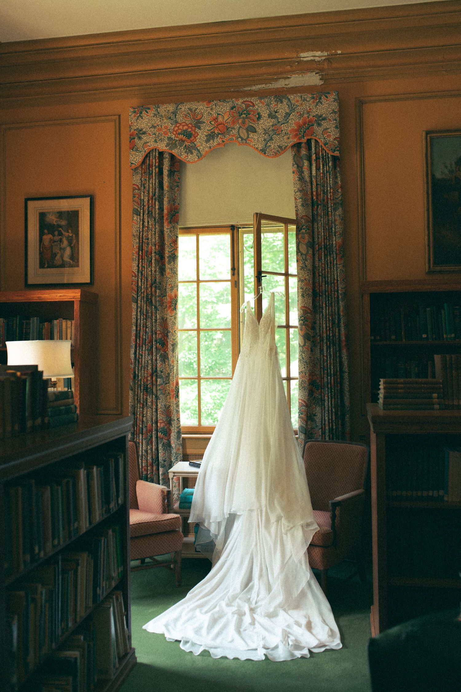 A flowing white wedding dress hangs in front of a window framed by floral curtains in a library setting.