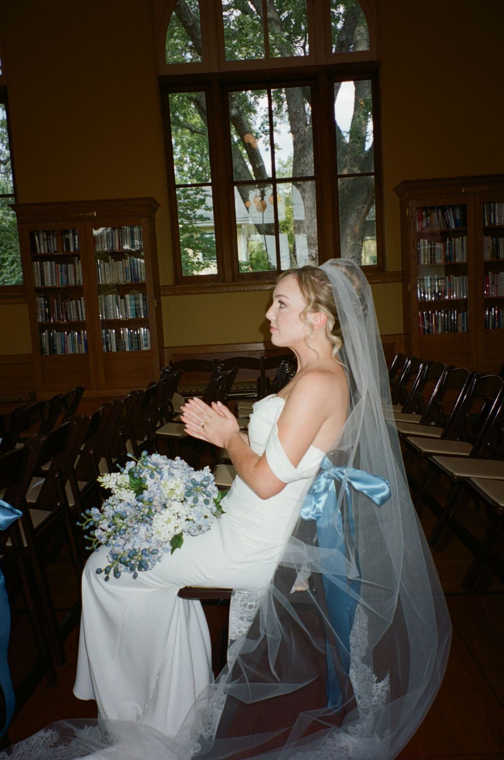 Bride seated in library setting with flowing veil and blue floral bouquet.