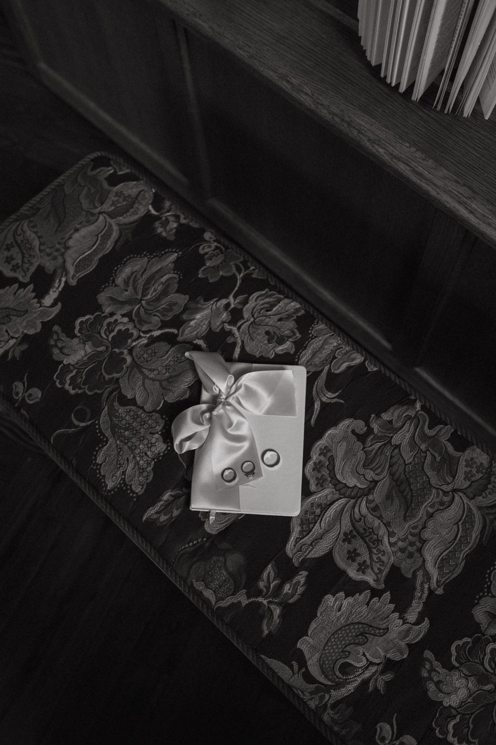 Wedding rings tied with white ribbon on dark floral upholstered bench.