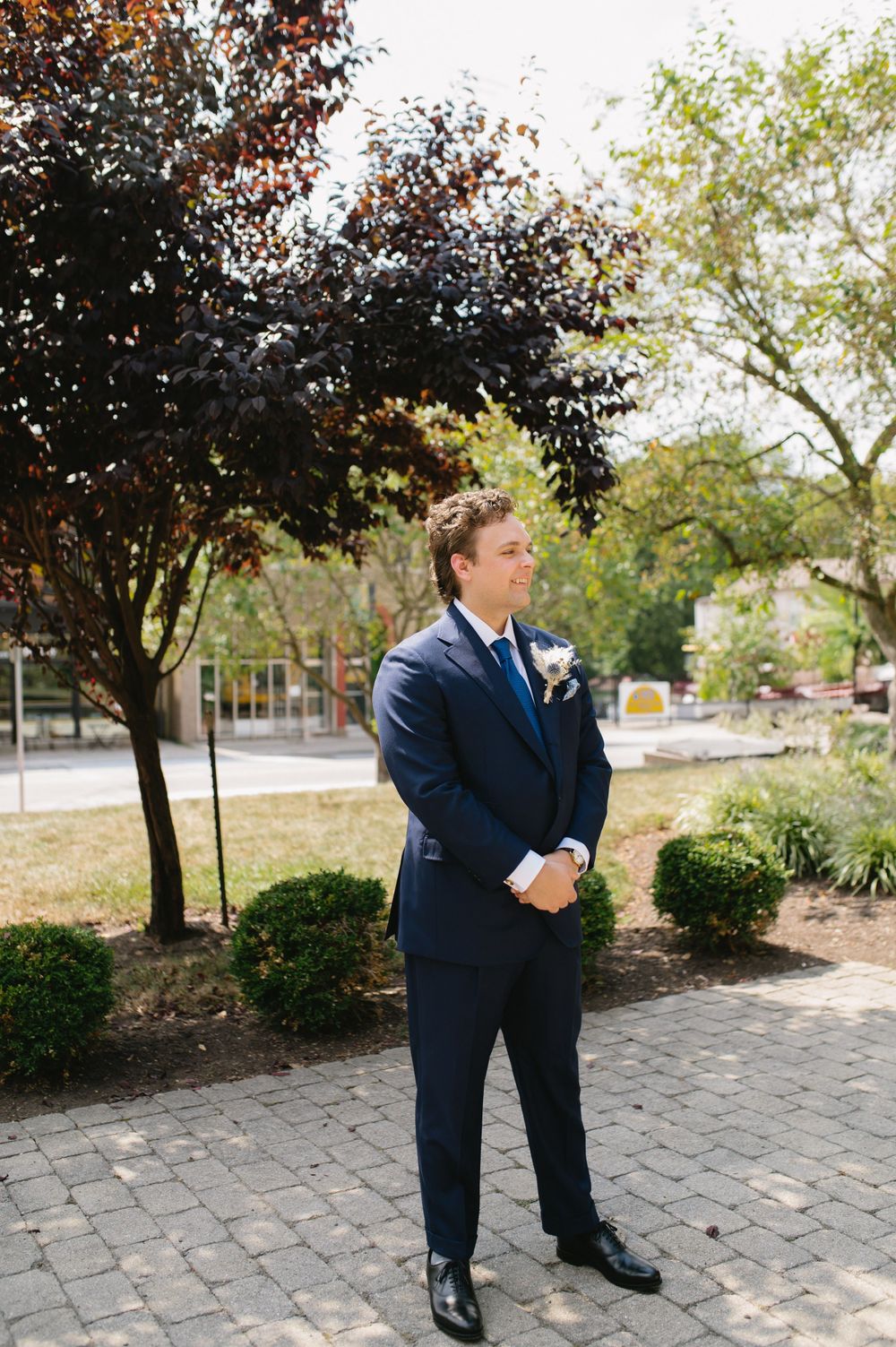 A groom in a navy suit stands outdoors on a brick path surrounded by autumn trees.