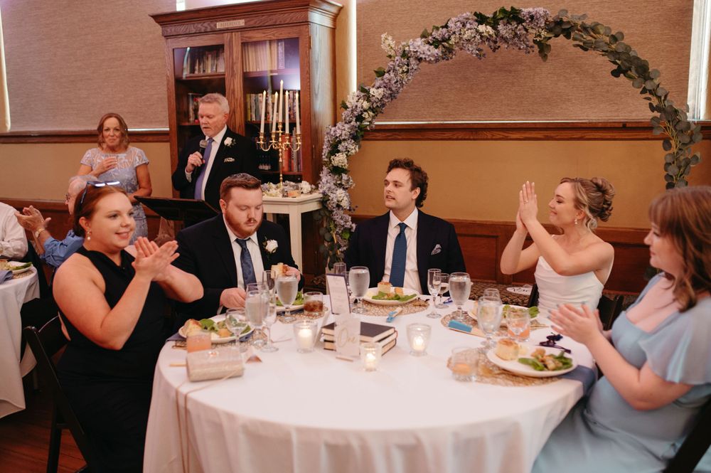 Wedding guests share laughter around a decorated table under a floral arch.