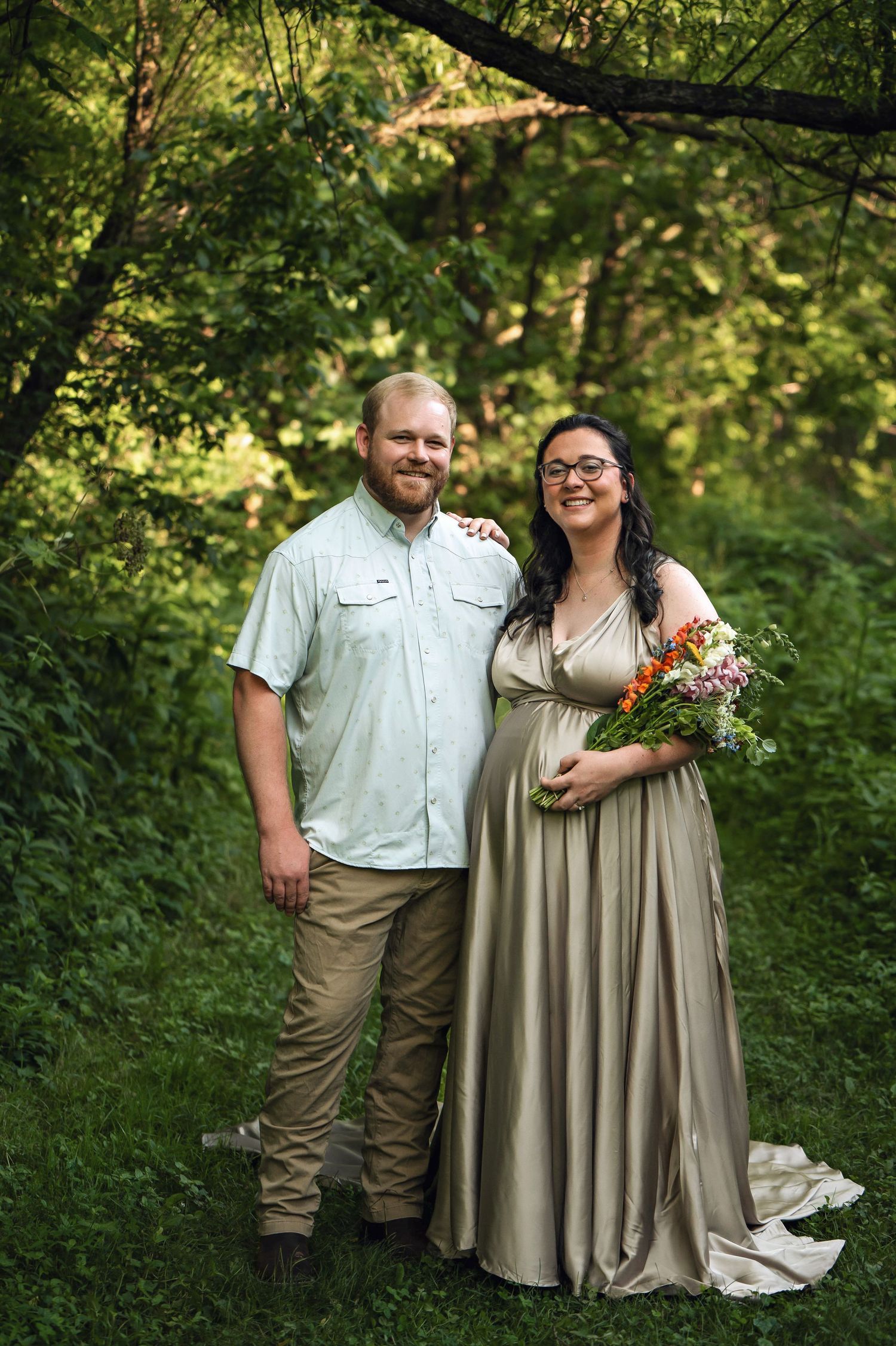 a maternity mom with a gold gown with a floral bouquet and her husband a wooded setting