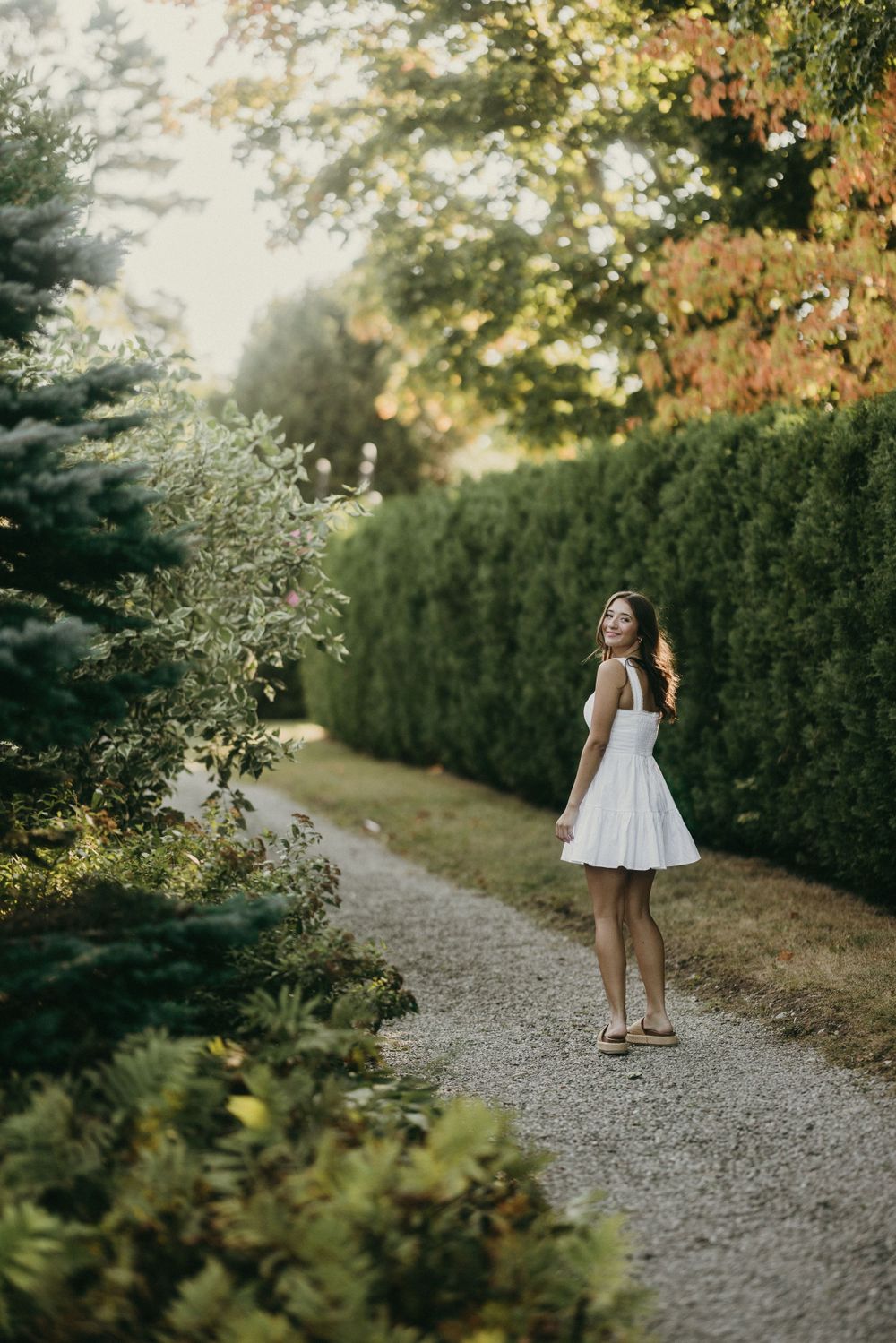 White dress on gravel path between green hedges during golden hour.