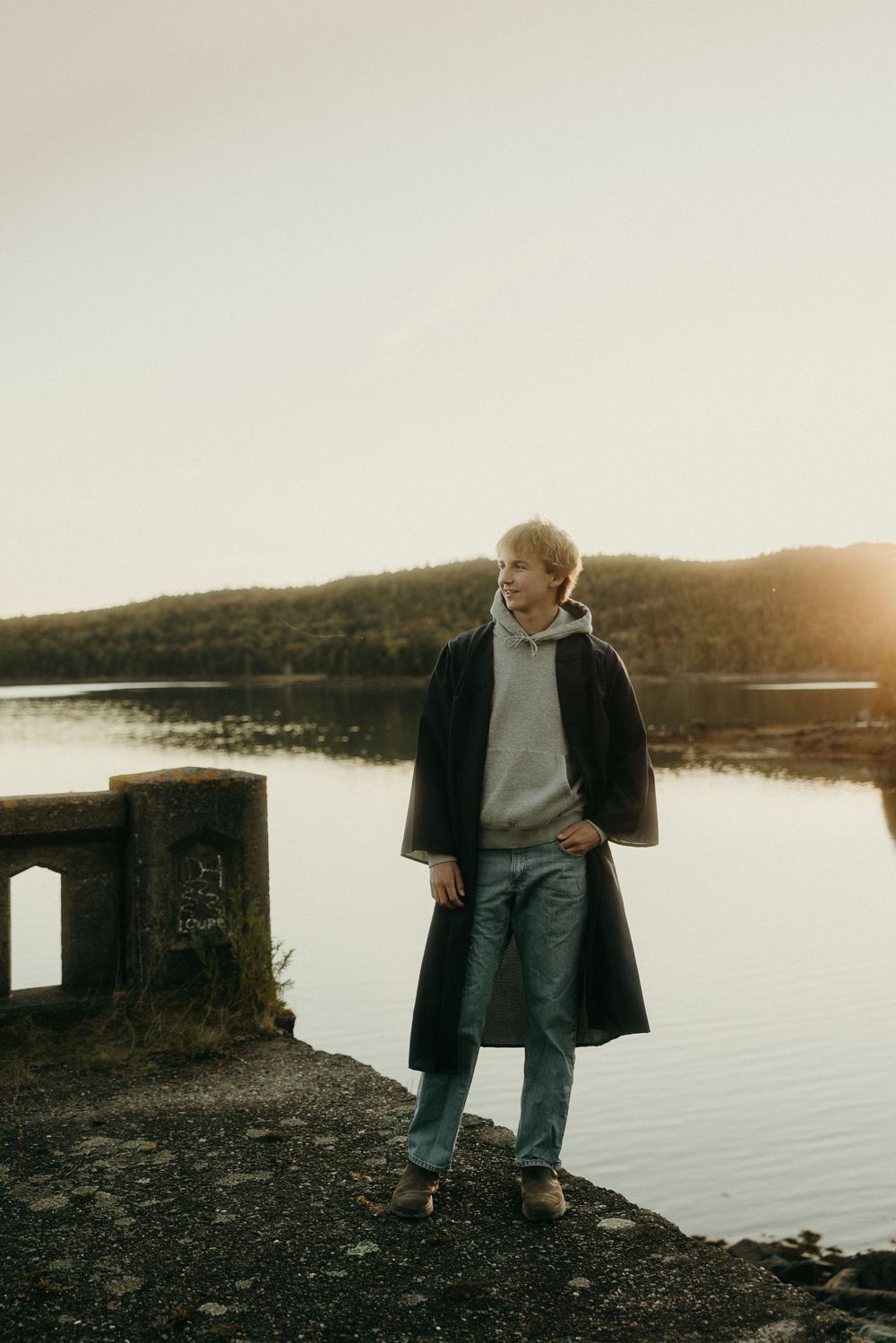 Figure sitting by a tranquil lake at dusk wearing a dark coat.