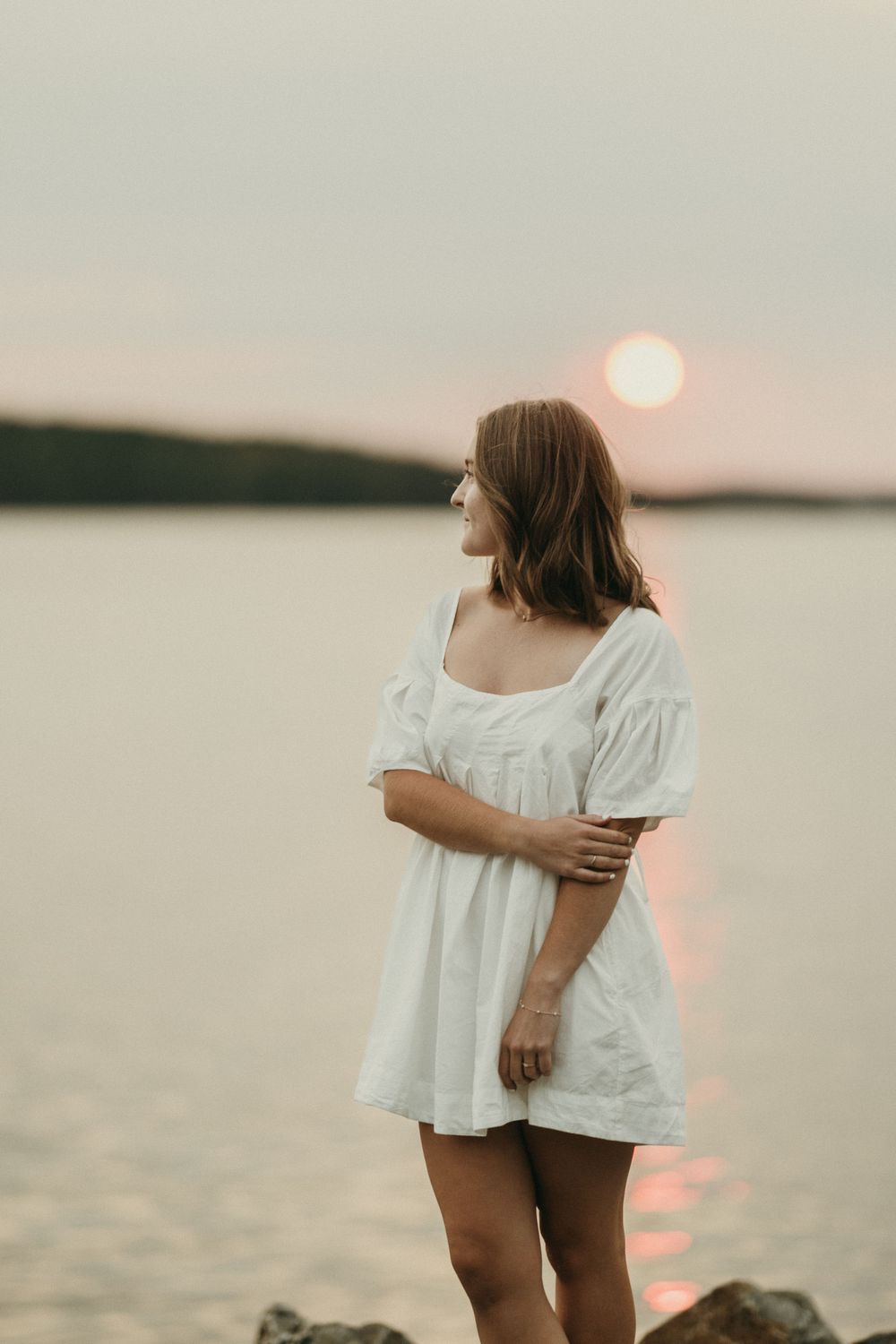 A person in a white top stands by a lake at sunset with a warm glow on the water.