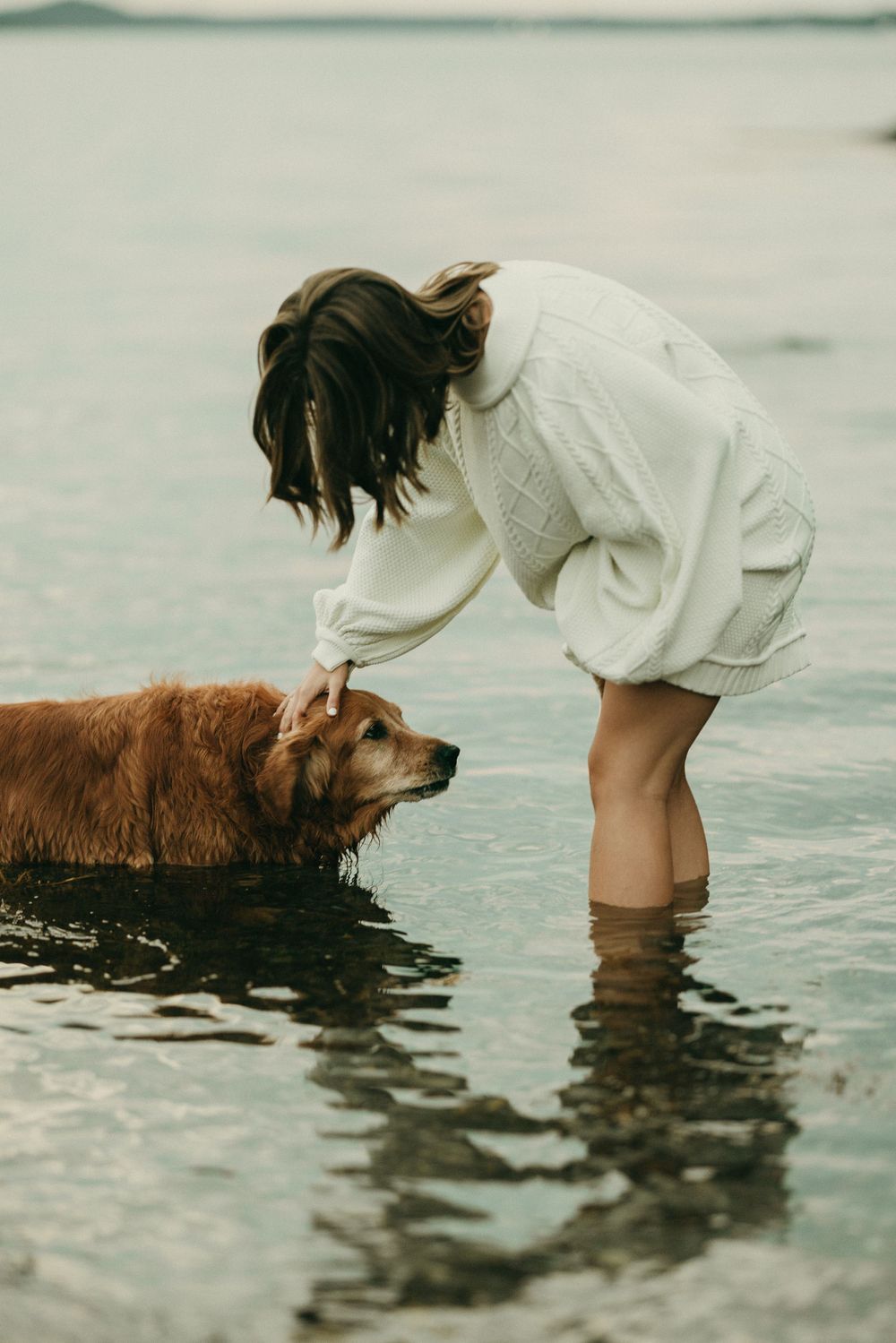 A person in a white dress bends down to pet their golden retriever in shallow water.