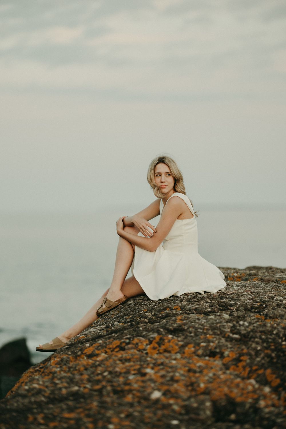 Person in white dress reclining on rocky coastal outcrop at dusk.