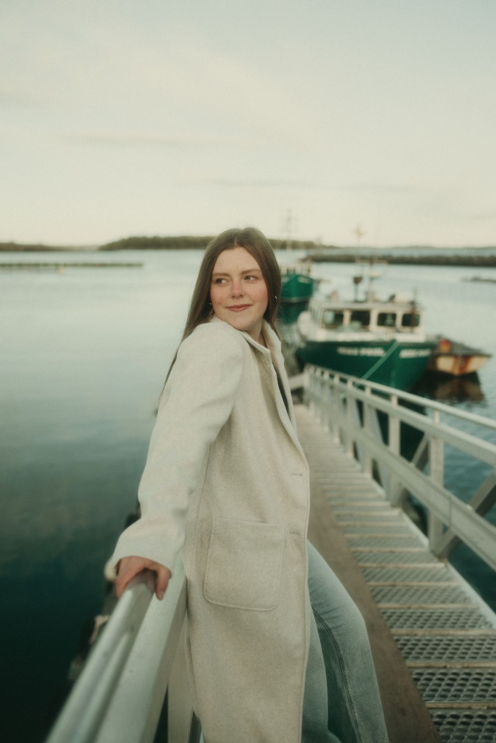 Someone in a cream sweater stands on a dock with boats and water in the background.