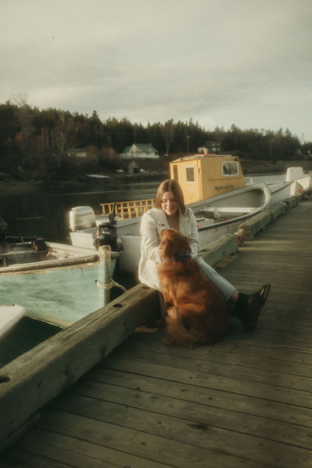 Person sitting with a dog on a wooden dock overlooking boats and water.