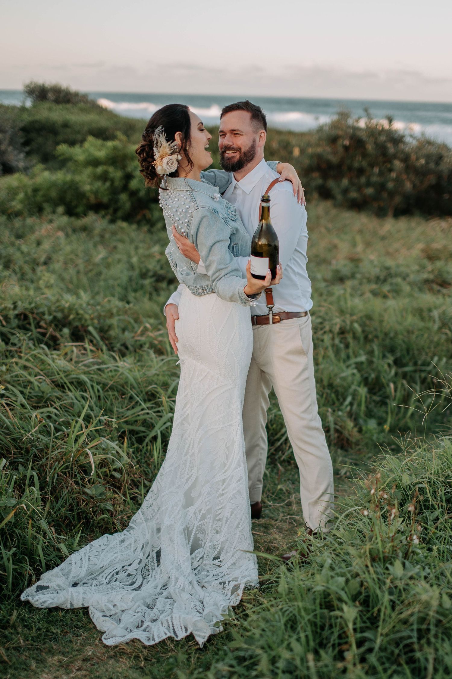 Couple in wedding attire sharing intimate moment on coastal hilltop with champagne.