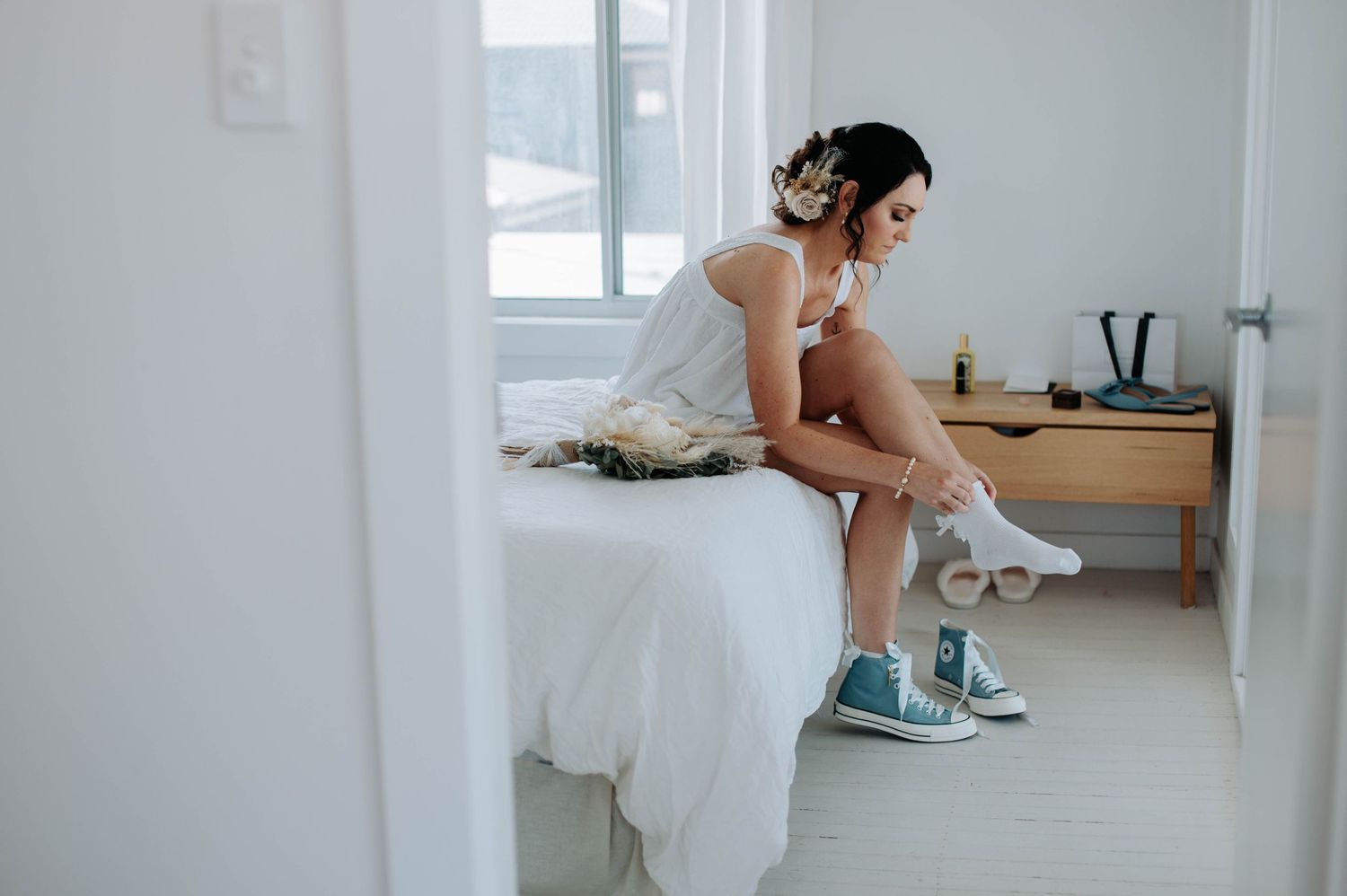 Person in wedding dress sitting on bed wearing blue sneakers.