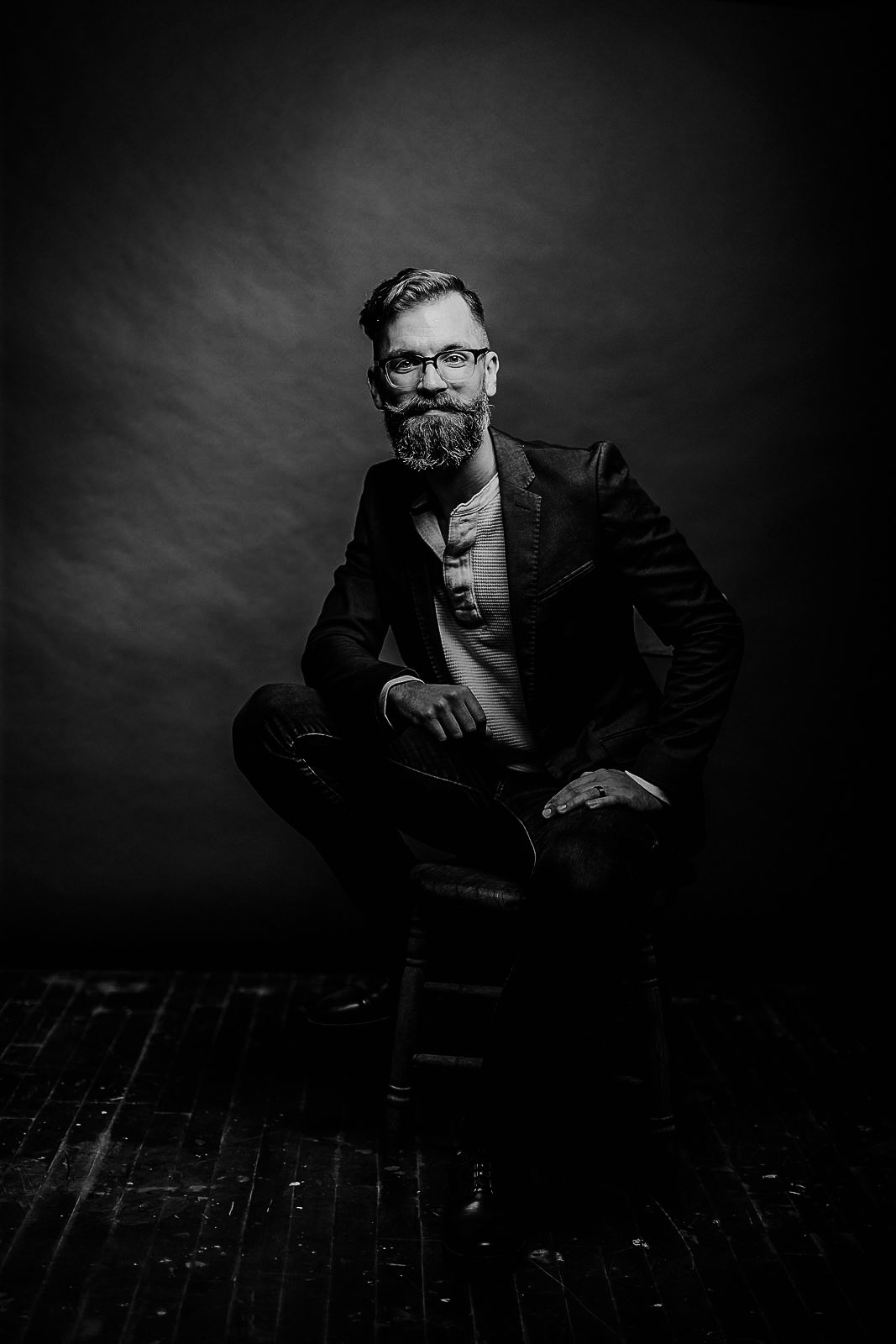 Moody low-key portrait of a seated figure in formal attire against dark background.