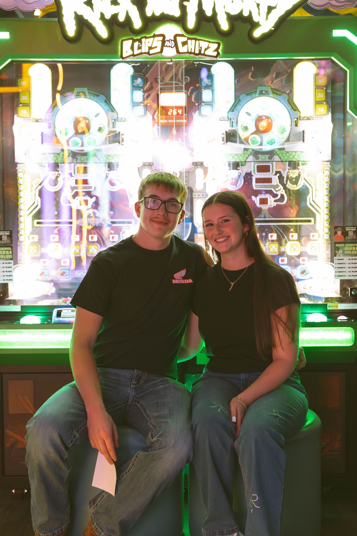 Two people sit together in front of a bright arcade game display with neon green lights and colorful graphics.