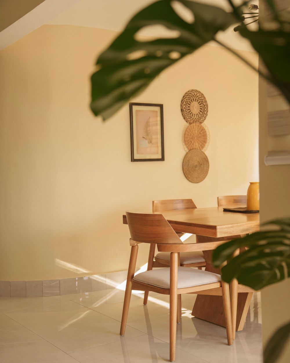 Modern dining area with wooden furniture and decorative woven wall art under a monstera plant's shadow.
