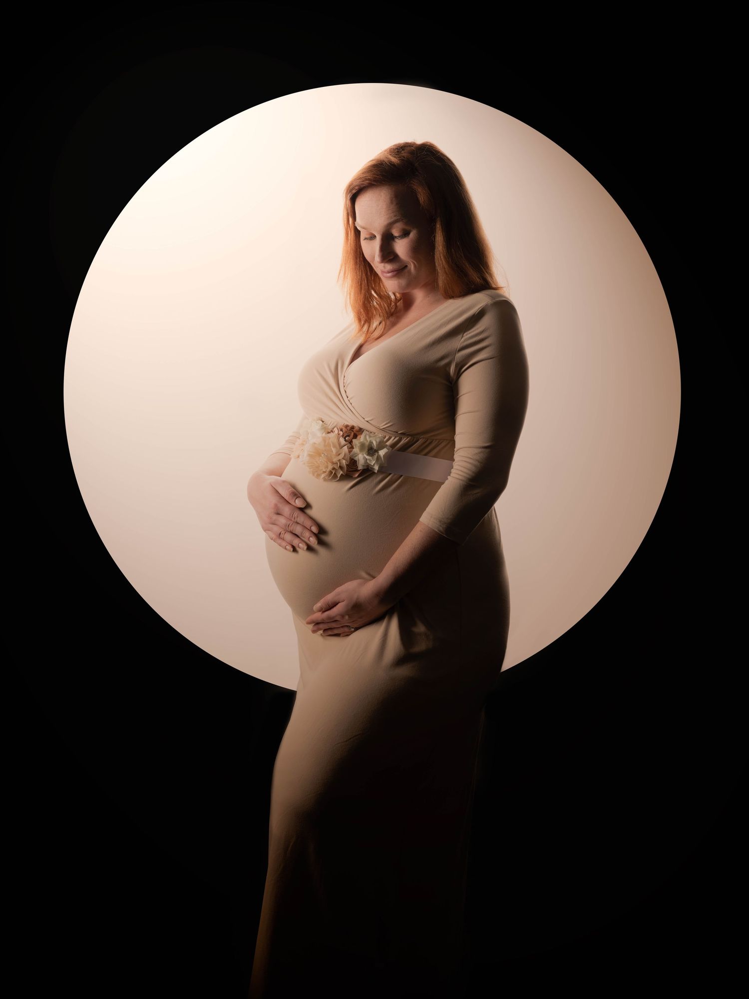 A pregnant woman in a beige dress poses against a dark background with dramatic circular lighting effect.