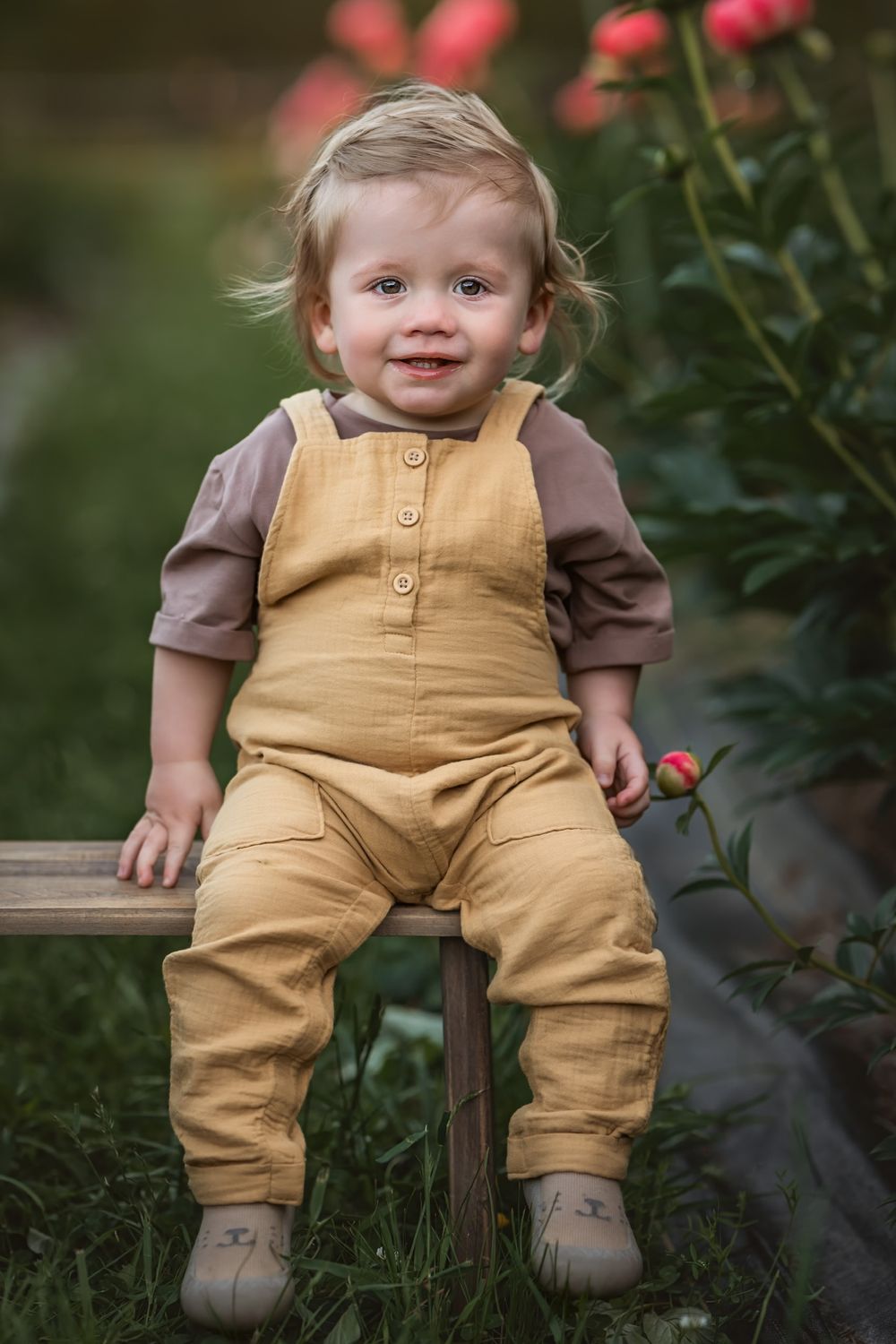 Small child wearing brown corduroy overalls sits on wooden bench near pink flowers in garden setting.