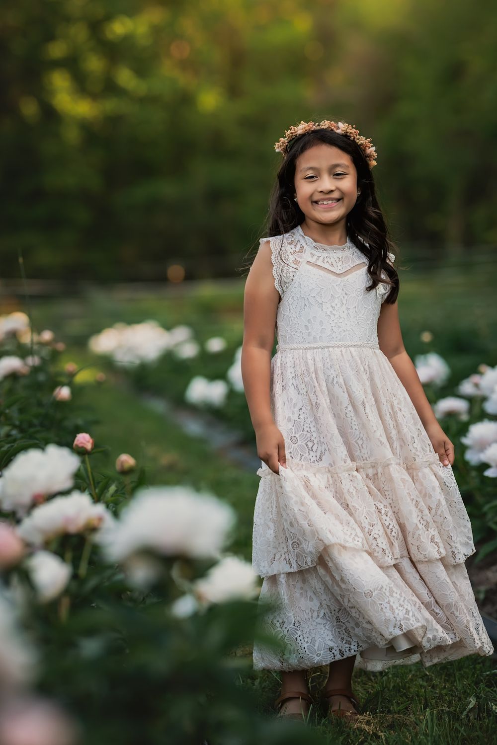 A young child wearing a white lace tiered dress and flower crown poses in a garden among blooming peonies.
