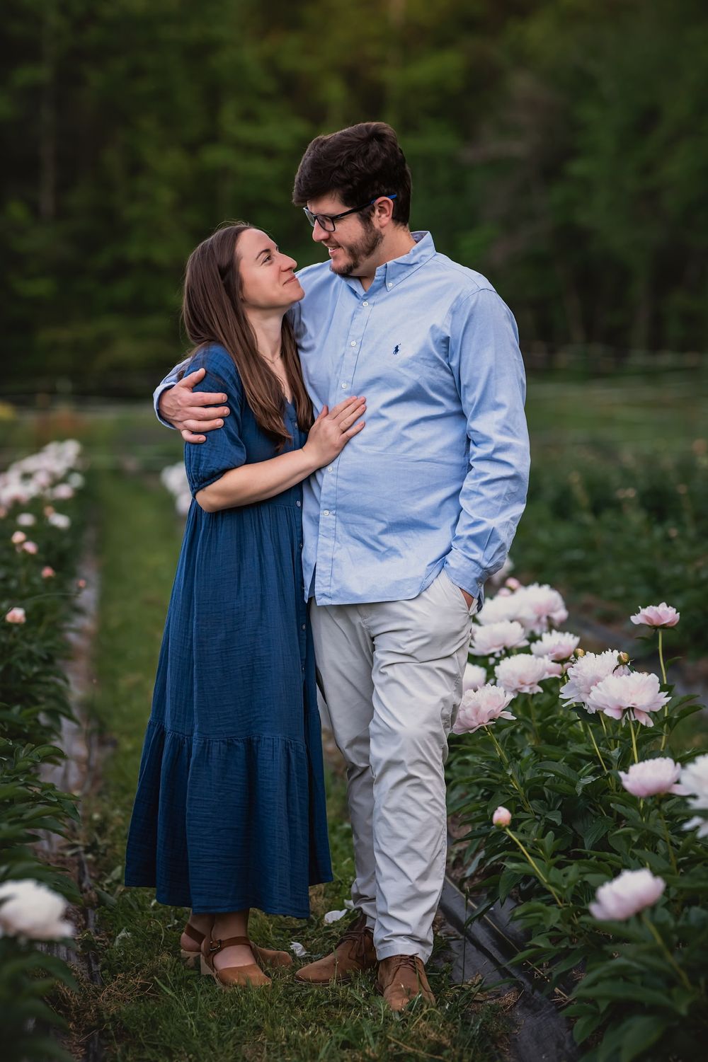 A couple embraces in a garden with blooming pink peonies, wearing blue and light colored casual attire.