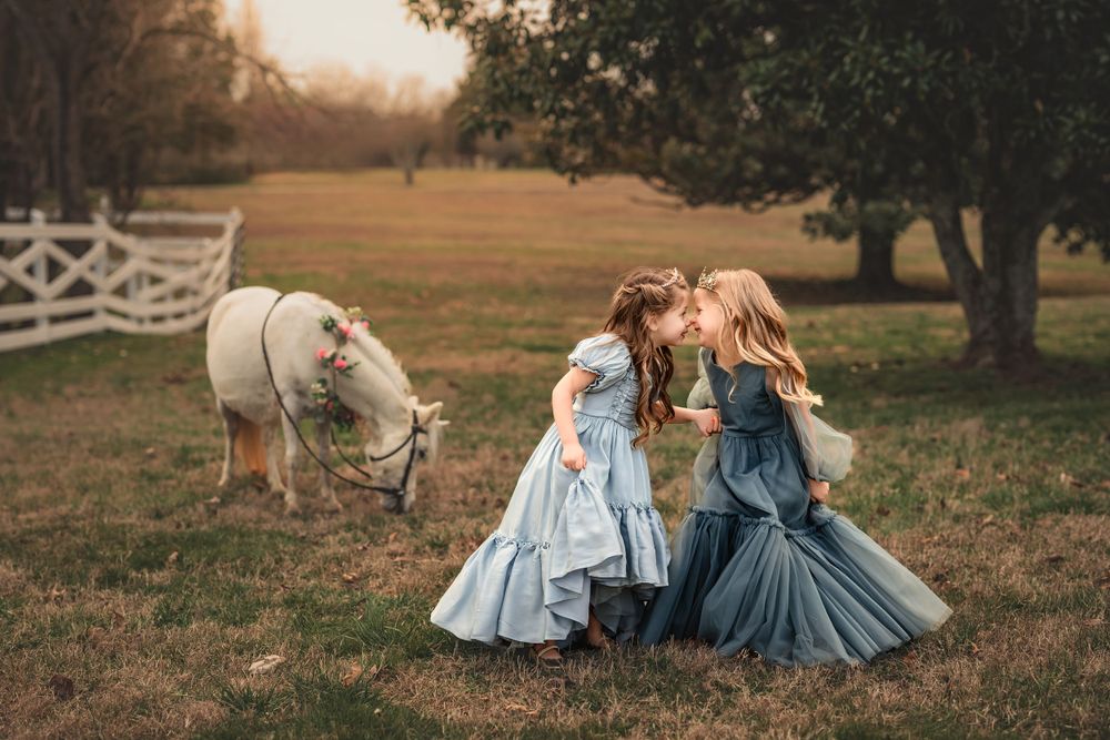 Two young children in flowing dresses share secrets in a pastoral field at sunset while a white horse grazes nearby.