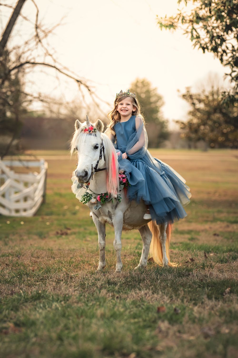Decorated white pony with floral wreath and rider in flowing blue dress stands in golden sunset light in pastoral field.