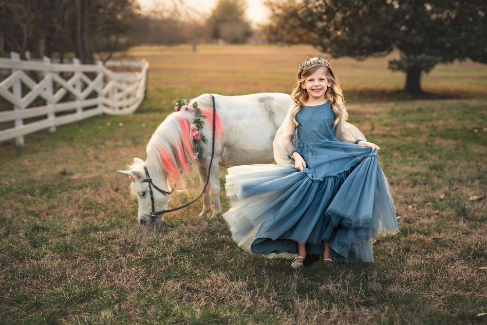 A whimsical scene of a young rider in a flowing blue dress posing with a decorated white pony at sunset.