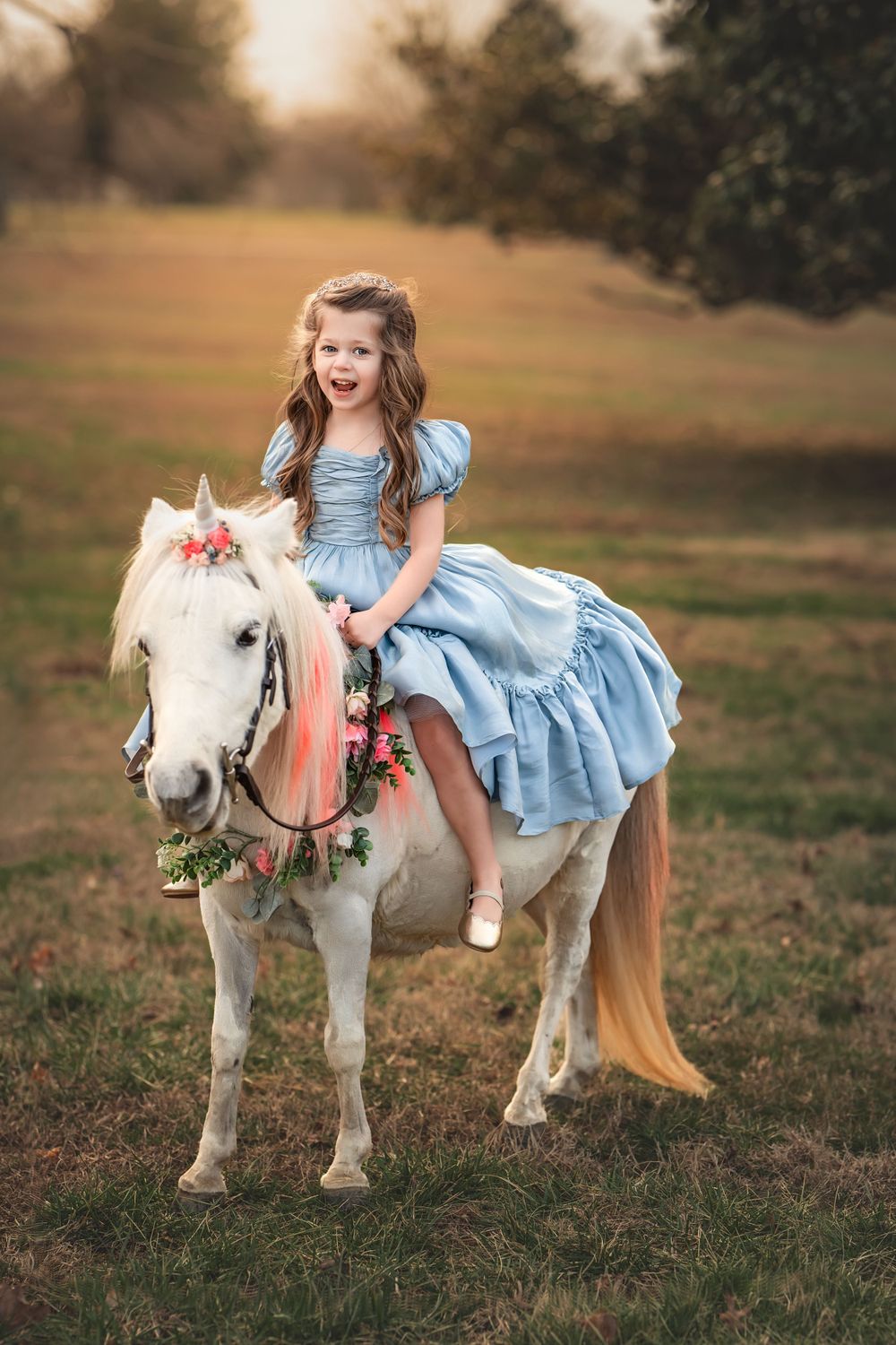 A small rider in a light blue dress sits atop a white pony wearing floral garland decorations at sunset.