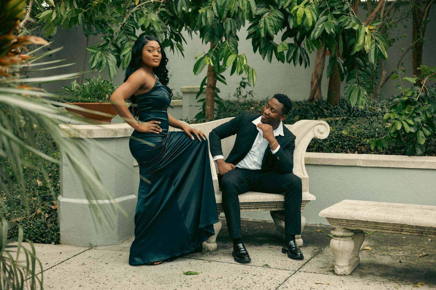Elegant couple poses in garden, woman in teal satin gown stands beside man seated in ornate white chair.