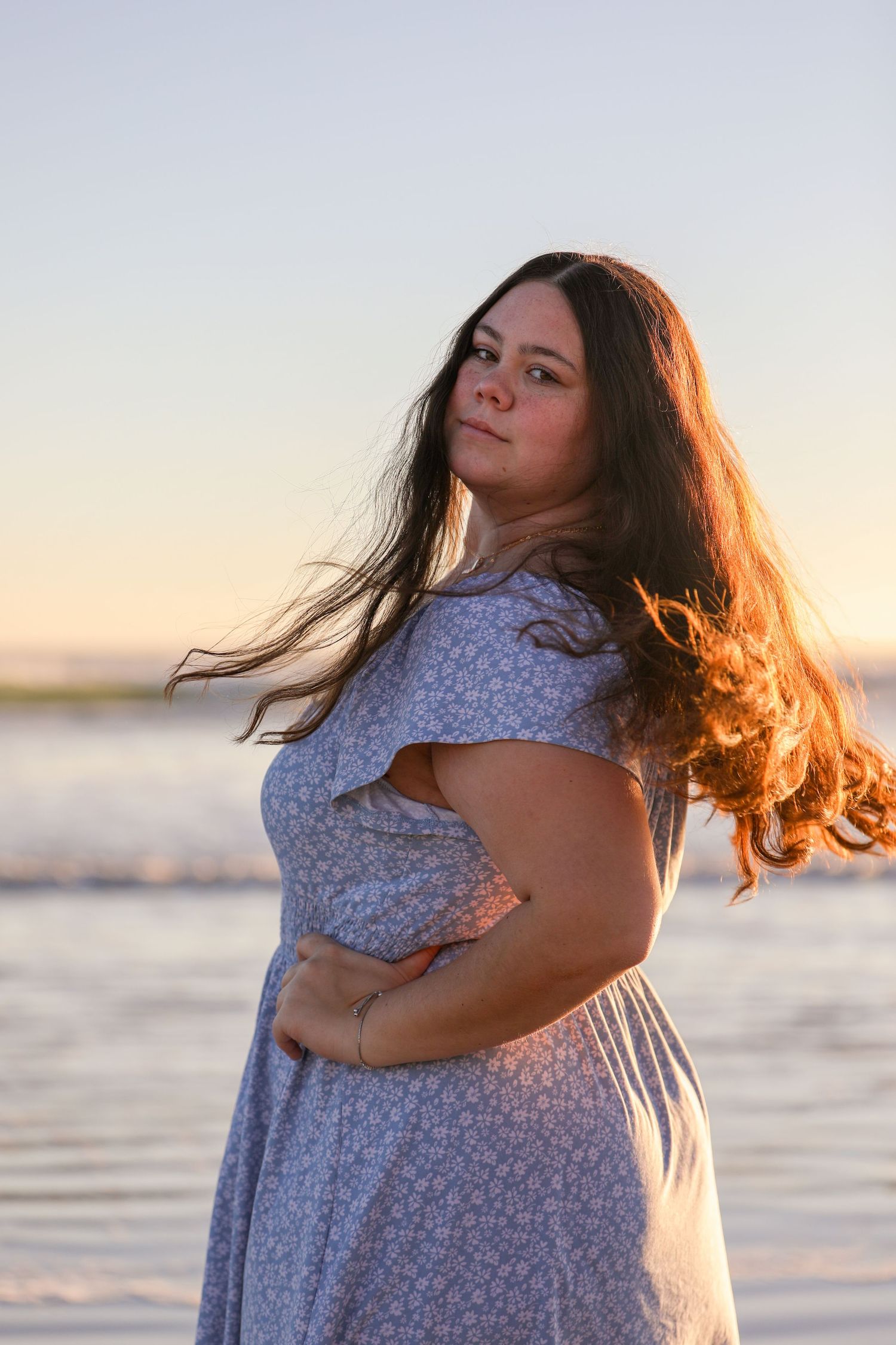 Person in a floral dress poses at sunset on the beach with hair flowing in the wind.