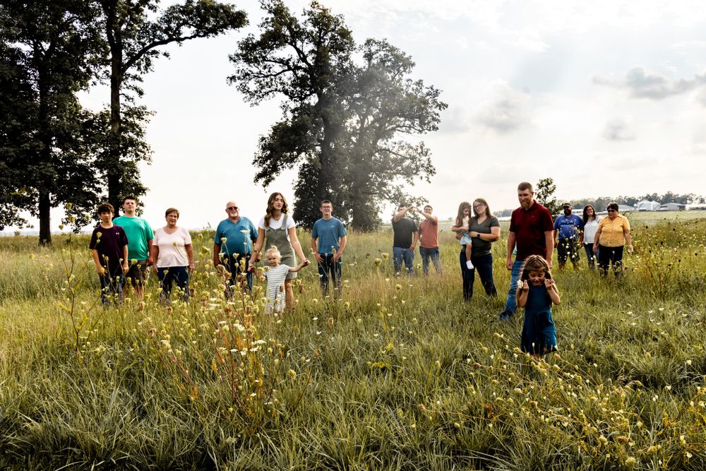 Group of people standing scattered in a sunny meadow with trees in the background.