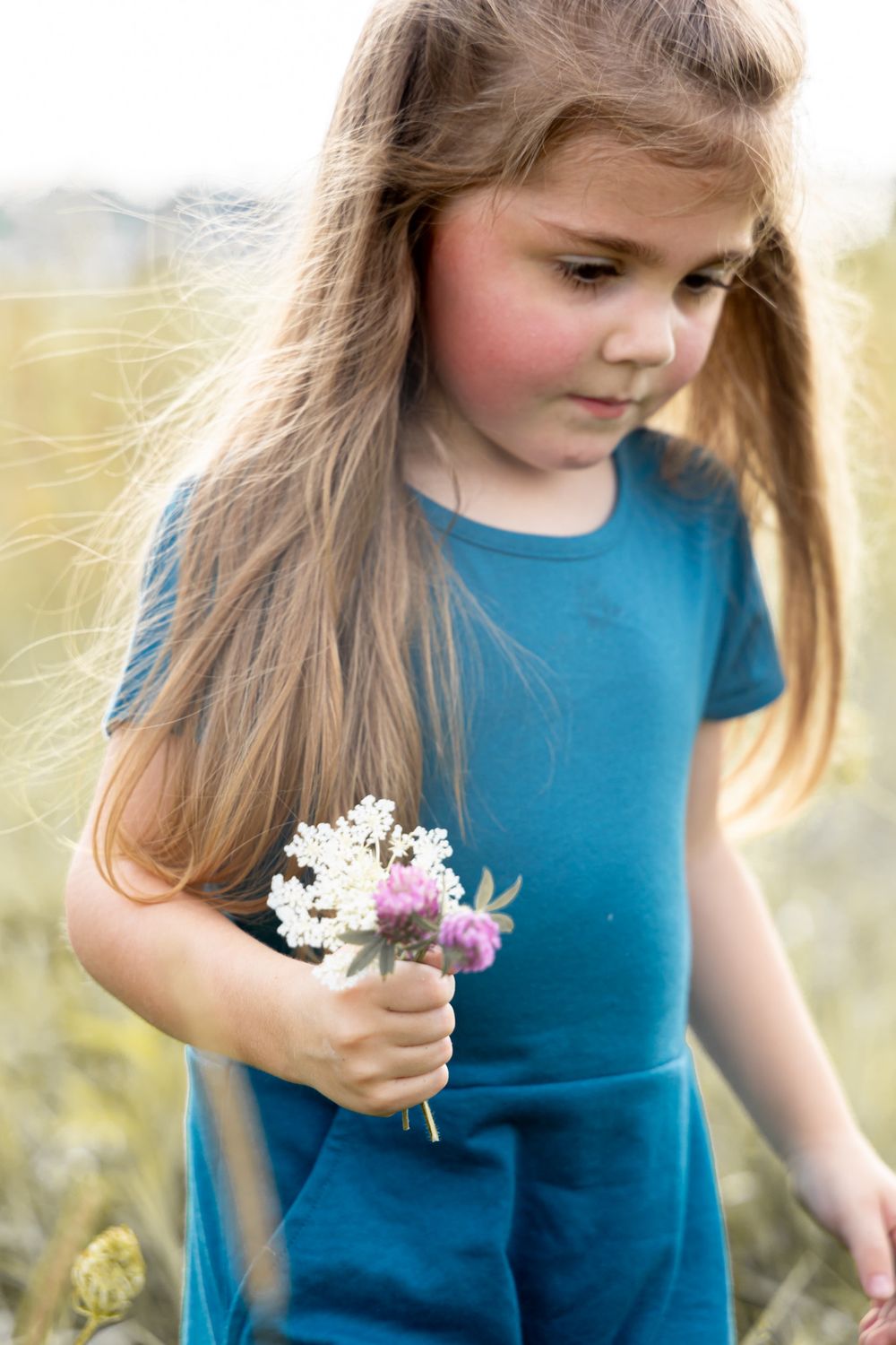 A young child in a blue dress holding wildflowers while standing in a summer field.