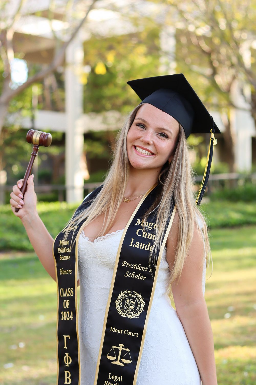 Graduate poses with graduation cap and honors stoles in outdoor garden setting.