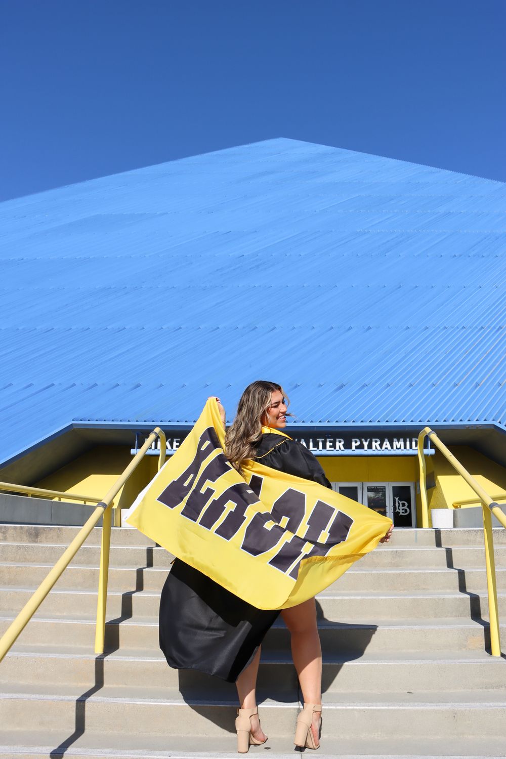 Graduate poses creatively on yellow architectural steps against blue sky background.