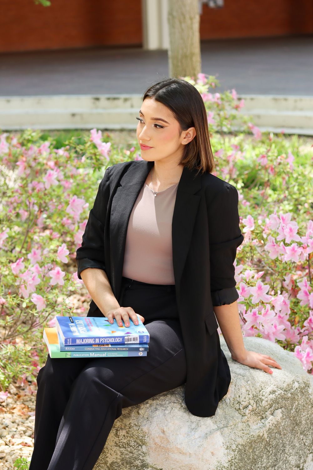 Student in black blazer poses with textbooks among pink flowers.