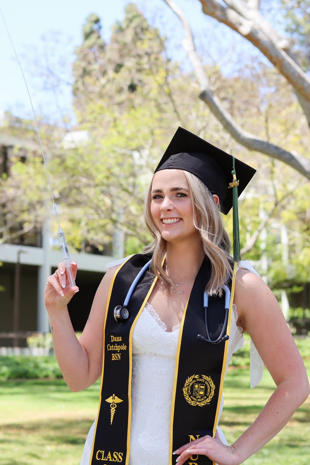 Graduate in white dress and black graduation stole poses with a stethoscope in an outdoor garden setting.