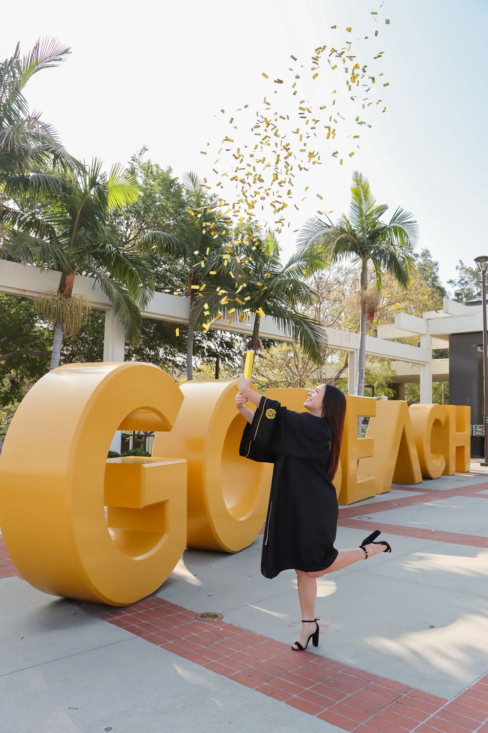 Graduate poses with large yellow CSULB letters during graduation celebration.