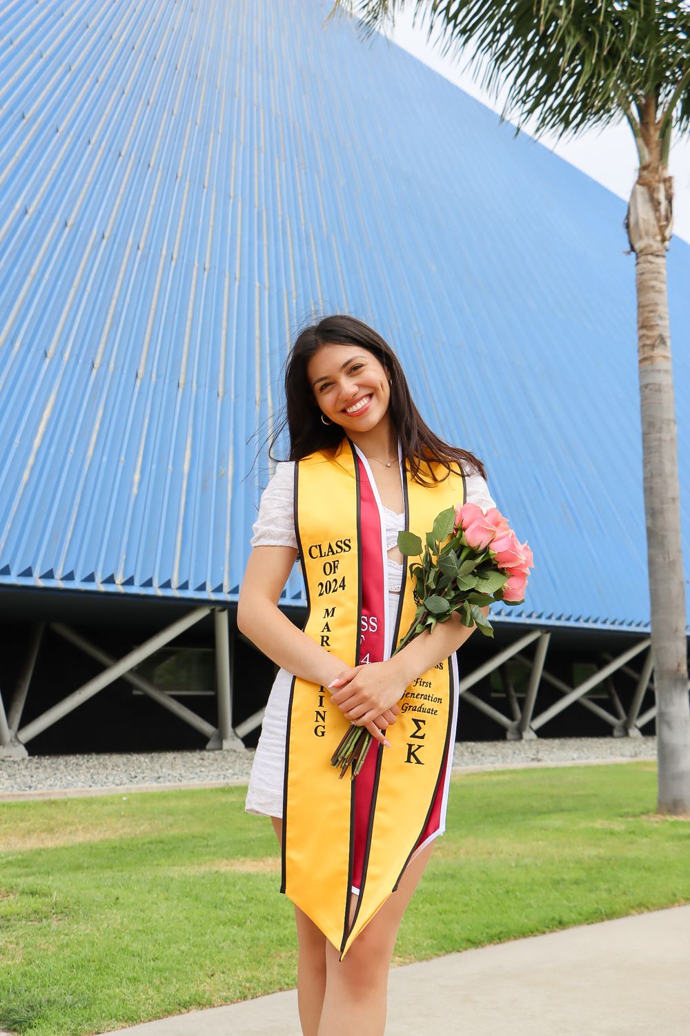 Graduate in white dress and gold stole poses with flowers near modern architecture.