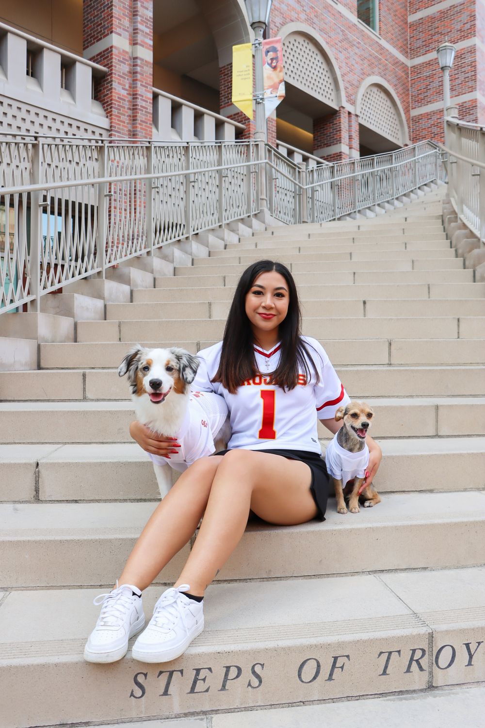 Graduate sits on Steps of Troy with two small dogs wearing matching outfits.