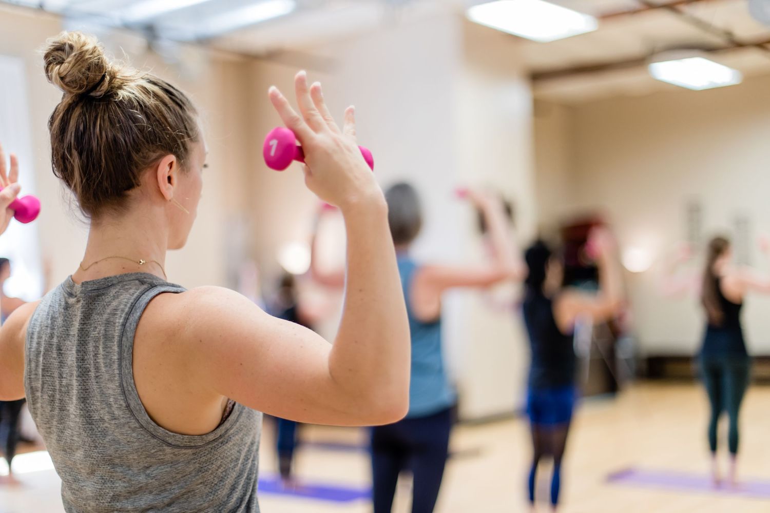 Workout class participants holding pink dumbbells during an exercise session in a bright fitness studio.