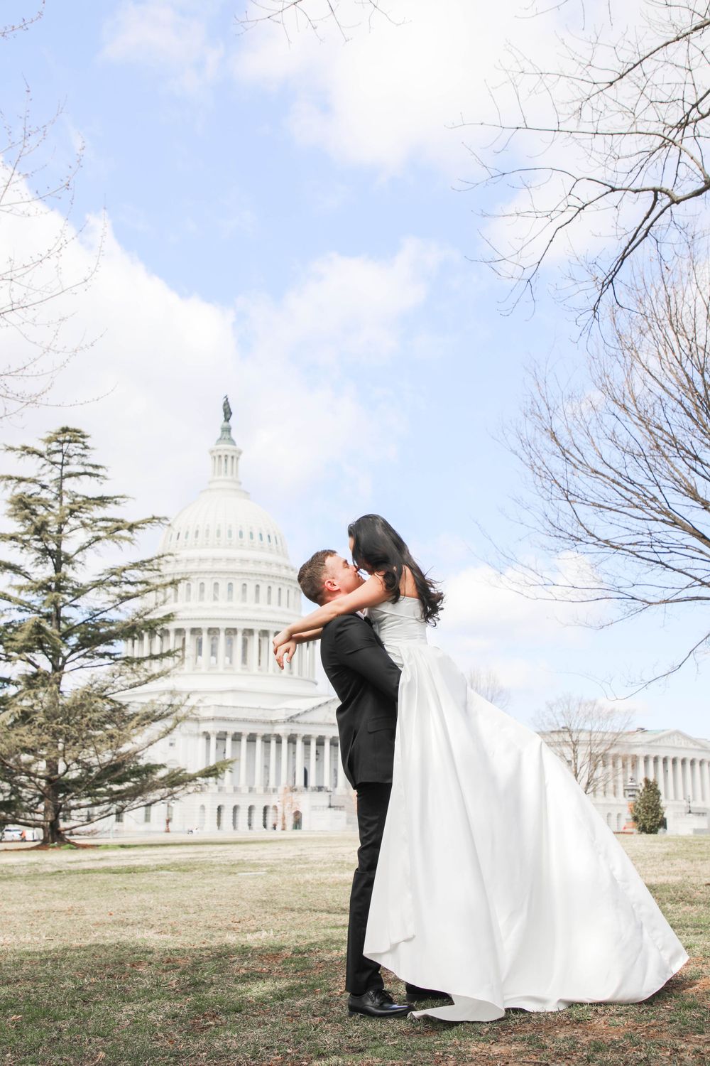 A couple in wedding attire embrace in front of the US Capitol Building on a sunny winter day.
