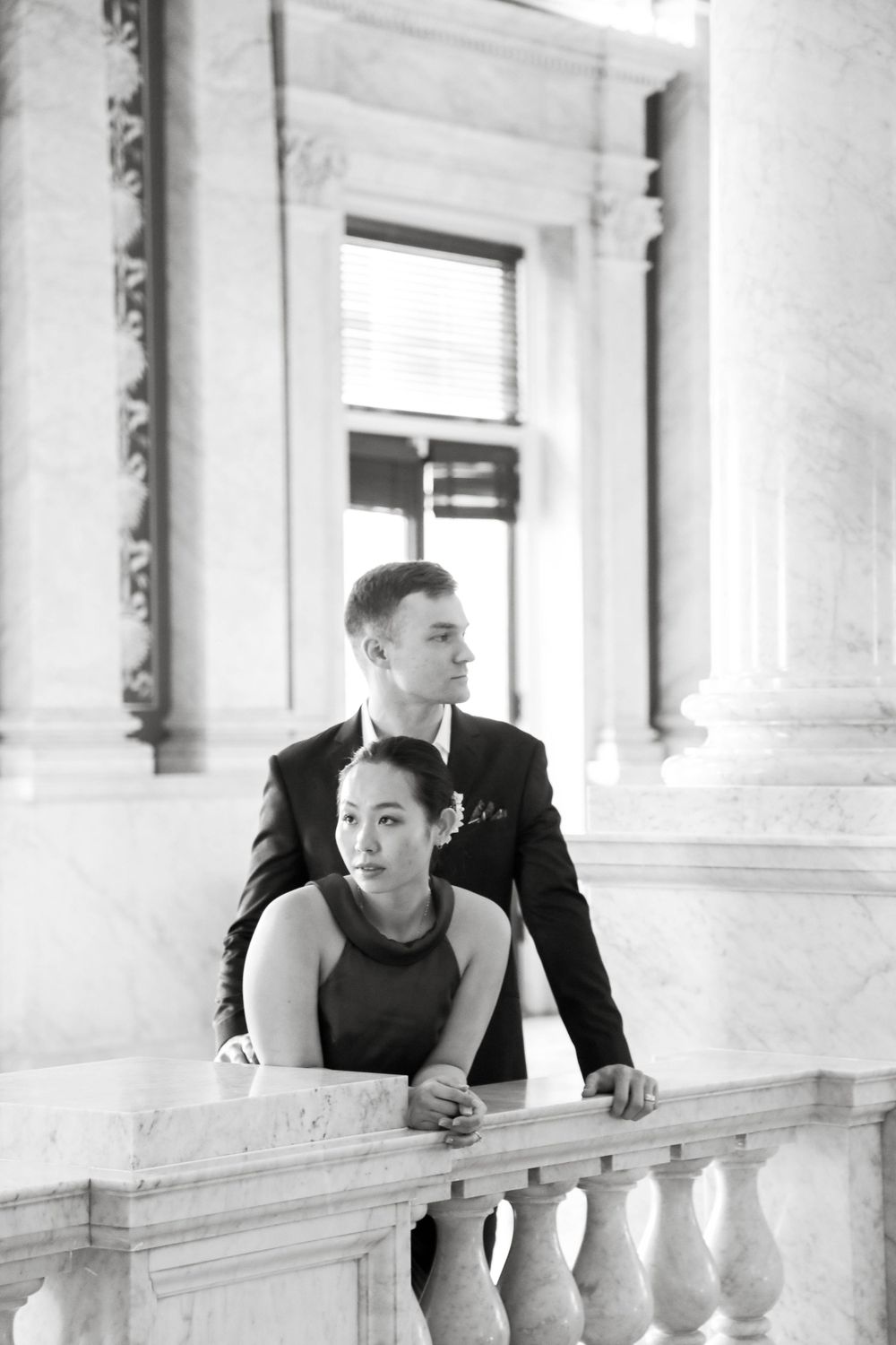A couple in formal attire poses together on an elegant white marble balcony in a black and white photograph.