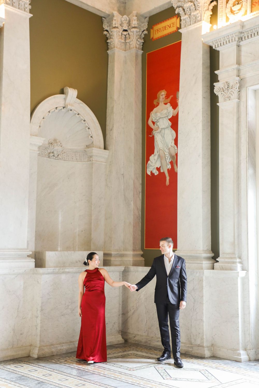 Elegant couple poses in formal attire inside ornate building with classical columns and red wall panel with white statue.