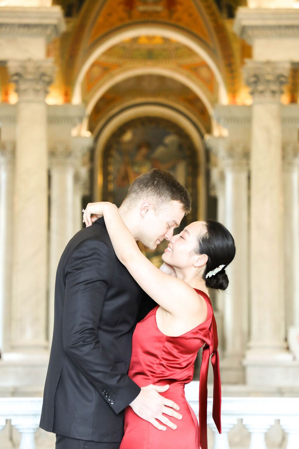 A couple in formal attire share a romantic embrace beneath ornate arched columns of a classical building.