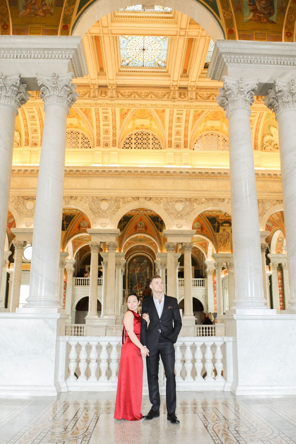 Elegant couple stands in formal attire beneath ornate gold and white architectural details at the Library of Congress.