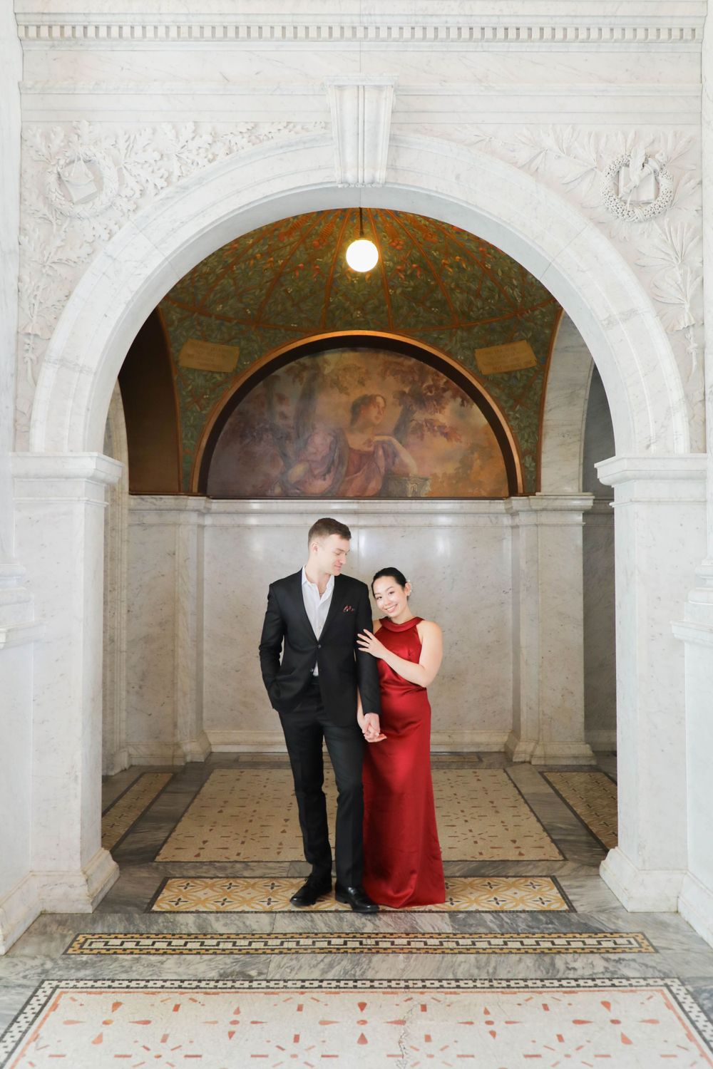 A couple in formal attire stands beneath an ornate archway with decorative ceiling and mural in a historic building.