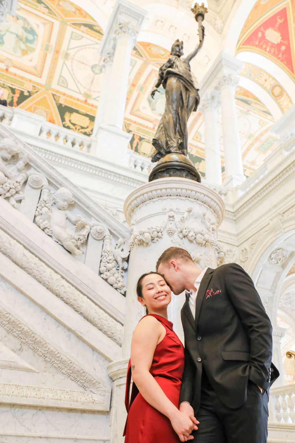 Couple in formal attire poses together on an elegant marble staircase beneath an ornate neoclassical statue and ceiling.