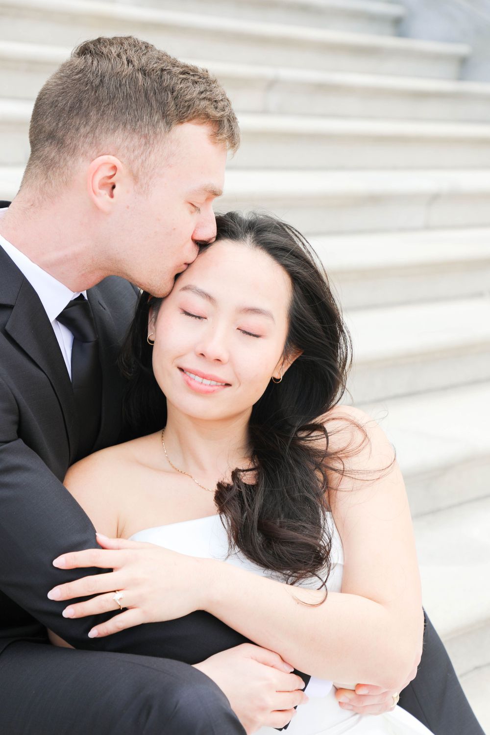 A tender wedding moment captured on white stone steps as a couple embraces in formal attire.