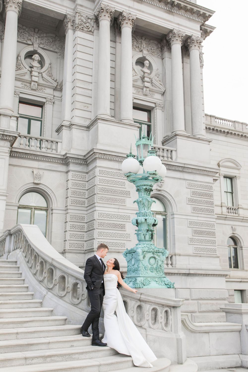 A couple stands on grand marble steps of a neoclassical building with ornate turquoise lampposts and architectural details.
